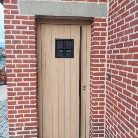 Houten deur inkom naturel met zwart raampje en zwart handvat geplaatst door Winfera